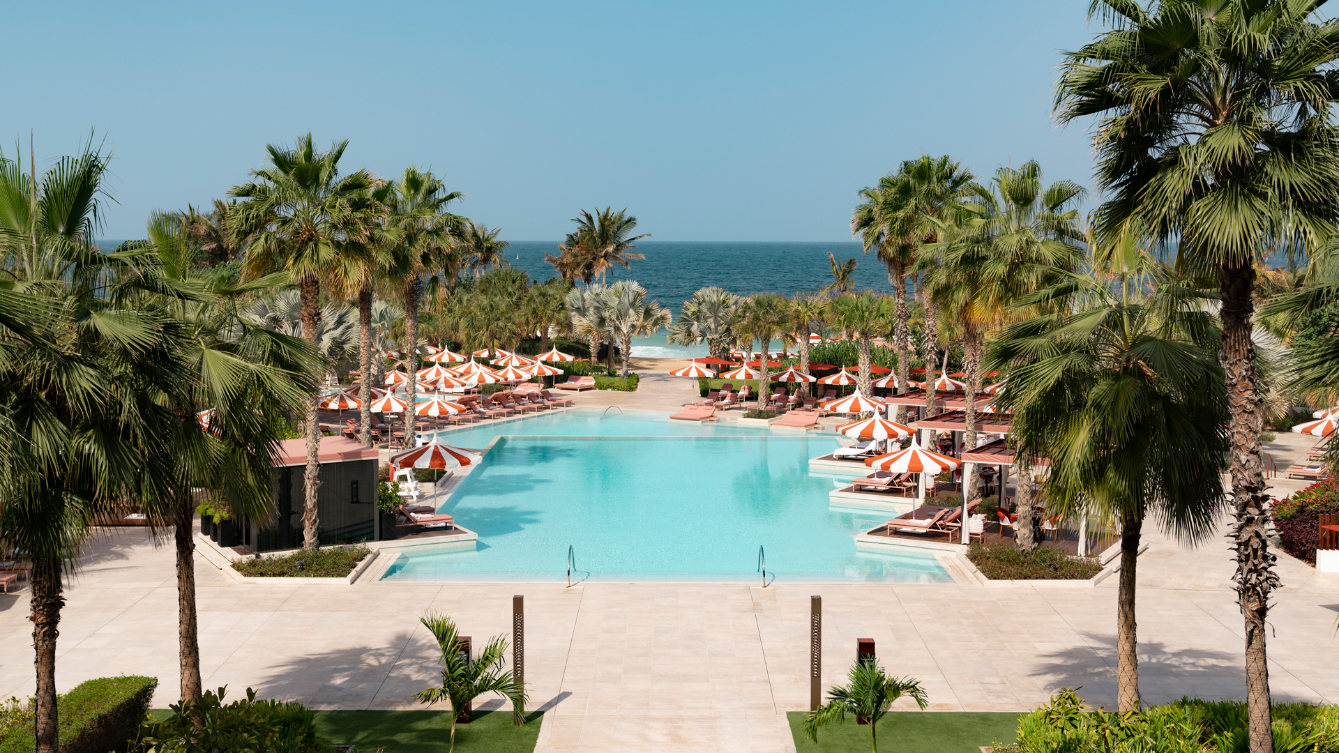 A stunning resort pool surrounded by lush palm trees and lined with elegant sun loungers and orange-and-white striped umbrellas, overlooking the pristine beach and the calm blue ocean in the background.